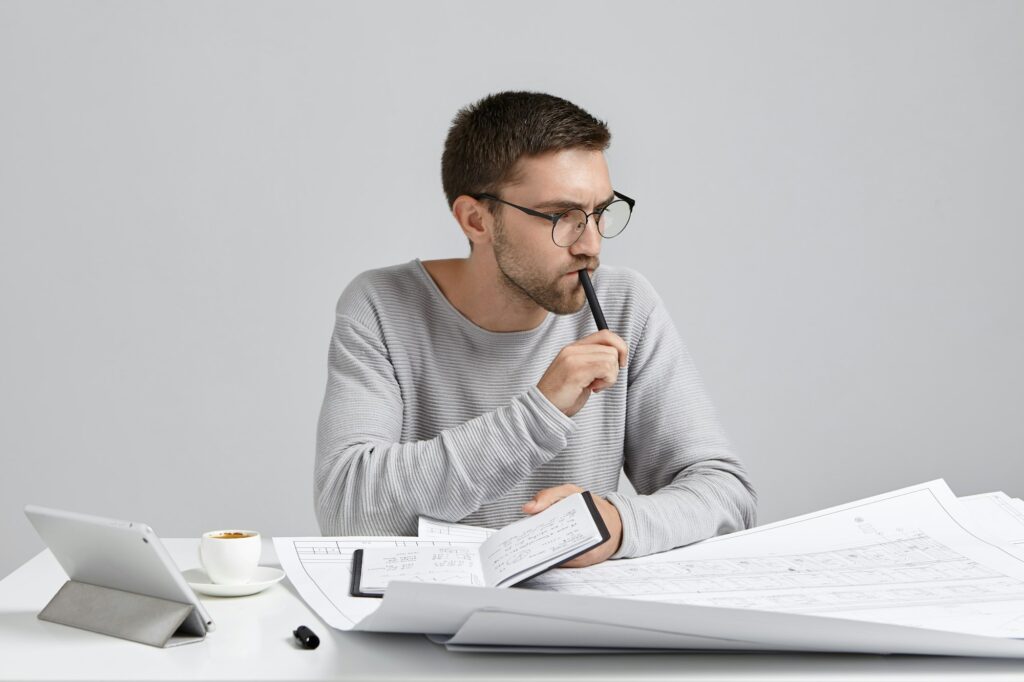 Serious pensive male engineer keeps pen and notebook in hands, plans meeting, look at drawings and s